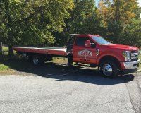 A bright red flatbed tow truck parked on an asphalt lot beside a leafy green tree on a sunny day.