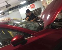 A person in a beanie and work uniform leans over the open hood of a bright red car in an auto repair shop.