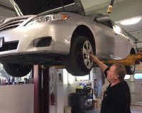 A mechanic in a garage inspects a silver car lifted on a hydraulic hoist.