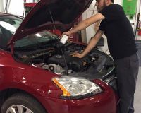 A person in a black shirt pours liquid from a plastic bottle into the engine bay of a red car in a garage.