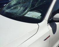 A white car with a shattered windshield and a parking violation notice placed under the wiper.