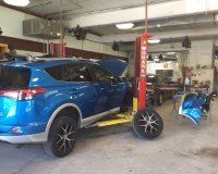 A bright blue SUV is raised on a red automotive lift inside a repair shop, with a detached car bumper resting nearby.