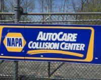 A blue and yellow NAPA AutoCare Collision Center sign mounted on a chain-link fence.