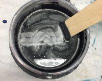 A wooden stirrer mixes silver and black swirling paint in a circular metal container.