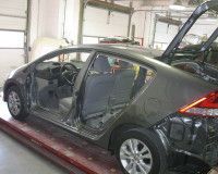 A dark gray sedan undergoing repairs inside an auto body shop, with its doors and rear bumper removed.