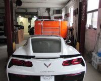 A white Chevrolet Corvette is parked inside an auto shop with an orange tool chest in the background.