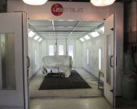 A view into an industrial car paint spray booth with a covered vehicle parked inside on a floor grate.