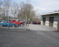 A blue car and a red truck parked behind a chain-link fence next to a gray industrial building on a sunny day.
