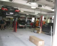 A bright auto repair shop interior with a car lifted on a hoist, a worker, and another vehicle parked on the floor.