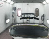 A black car bumper and spoiler prepared for painting inside a brightly lit industrial spray booth.