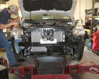 A mechanic works on the exposed engine and headlights of a car mounted on a lift inside an auto repair shop.