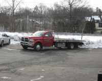 A red flatbed tow truck parked in a snowy, paved lot with trees in the background.