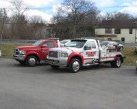 Two tow trucks, one red and one white with red accents, parked on a paved lot in front of a building and trees.