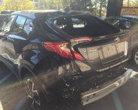 A damaged black Toyota C-HR parked in a lot, missing its rear bumper and with a shattered rear windshield.