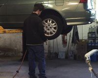 A mechanic works on the rear tire of a grey car raised on a hydraulic lift inside an auto repair shop.