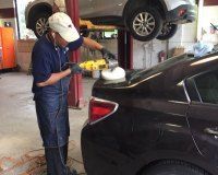 A person in a garage uses a handheld yellow power polisher to buff the trunk of a black car.