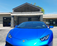 A bright blue Lamborghini parked in front of a commercial auto body shop building under a clear blue sky.