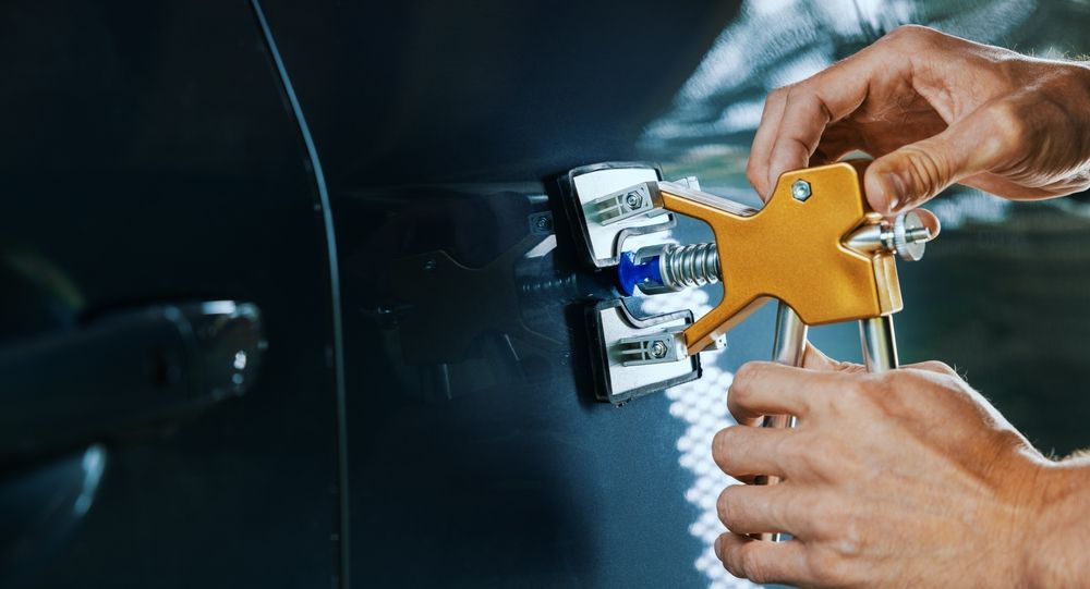 A person uses a gold-colored dent puller tool to repair a small dent on a dark vehicle door panel.