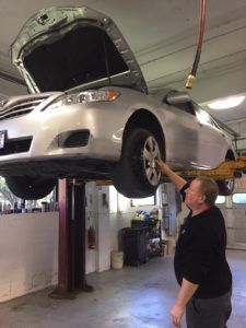 A mechanic inspects the front wheel of a silver sedan raised on a hydraulic lift in a garage.