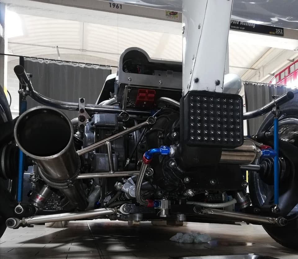 Vista frontale delle sospensioni posteriori e dello scarico a vista di un'auto da corsa in un garage, con componenti blu e un montante centrale del telaio.