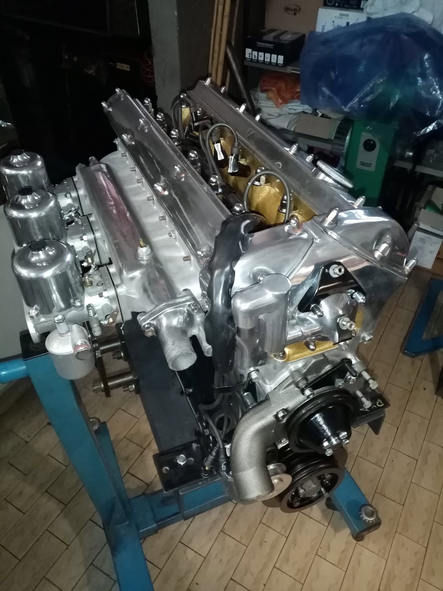 In un'officina, un blocco motore lucido a forma di V è appoggiato su un supporto blu, circondato da attrezzi e ricambi.