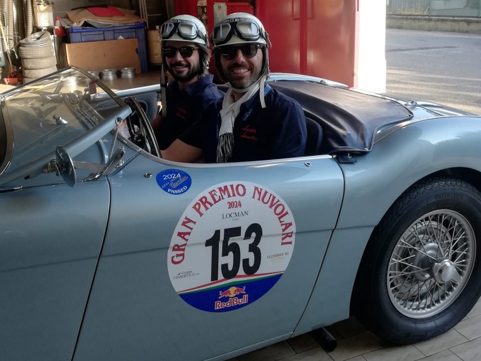 Due piloti con il casco siedono in un'auto da corsa d'epoca azzurra contrassegnata dal numero 153 sulla linea di partenza di un rally.