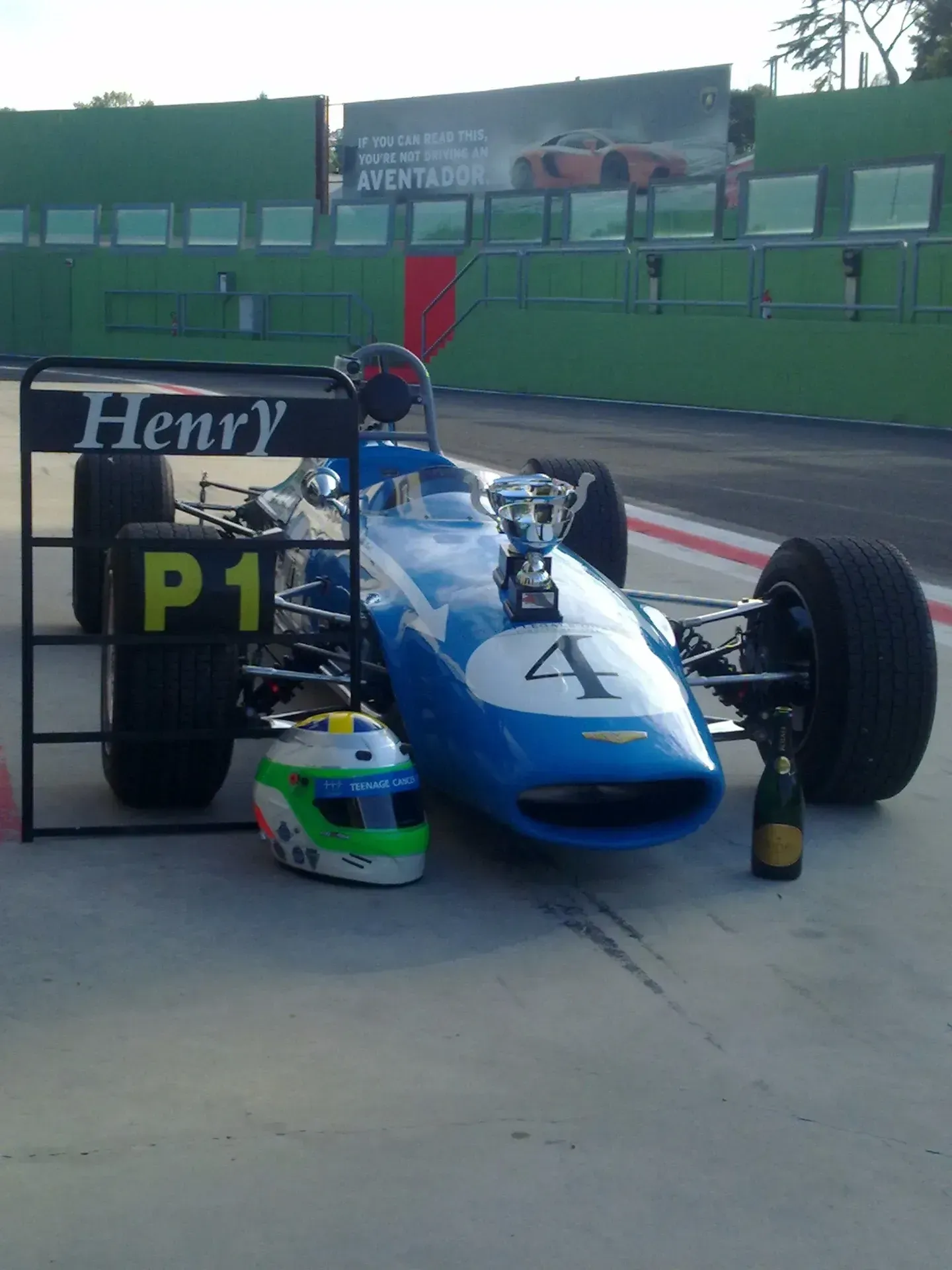 Macchina da corsa blu con il numero 4 e il cartello P1 su una pista, con casco e trofeo accanto.