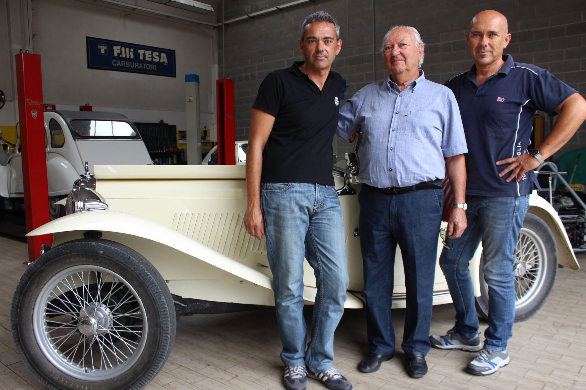 Tre uomini sono in piedi accanto a un'auto d'epoca color crema in un garage, e uno di loro tiene aperta la portiera.