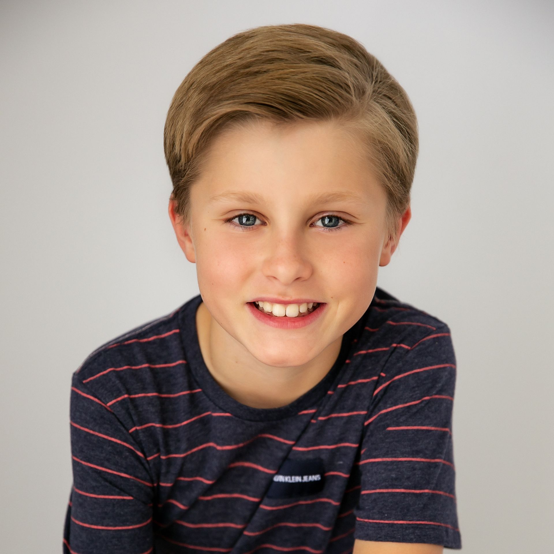 Smiling child with short blond hair wearing a dark striped shirt against a light gray background