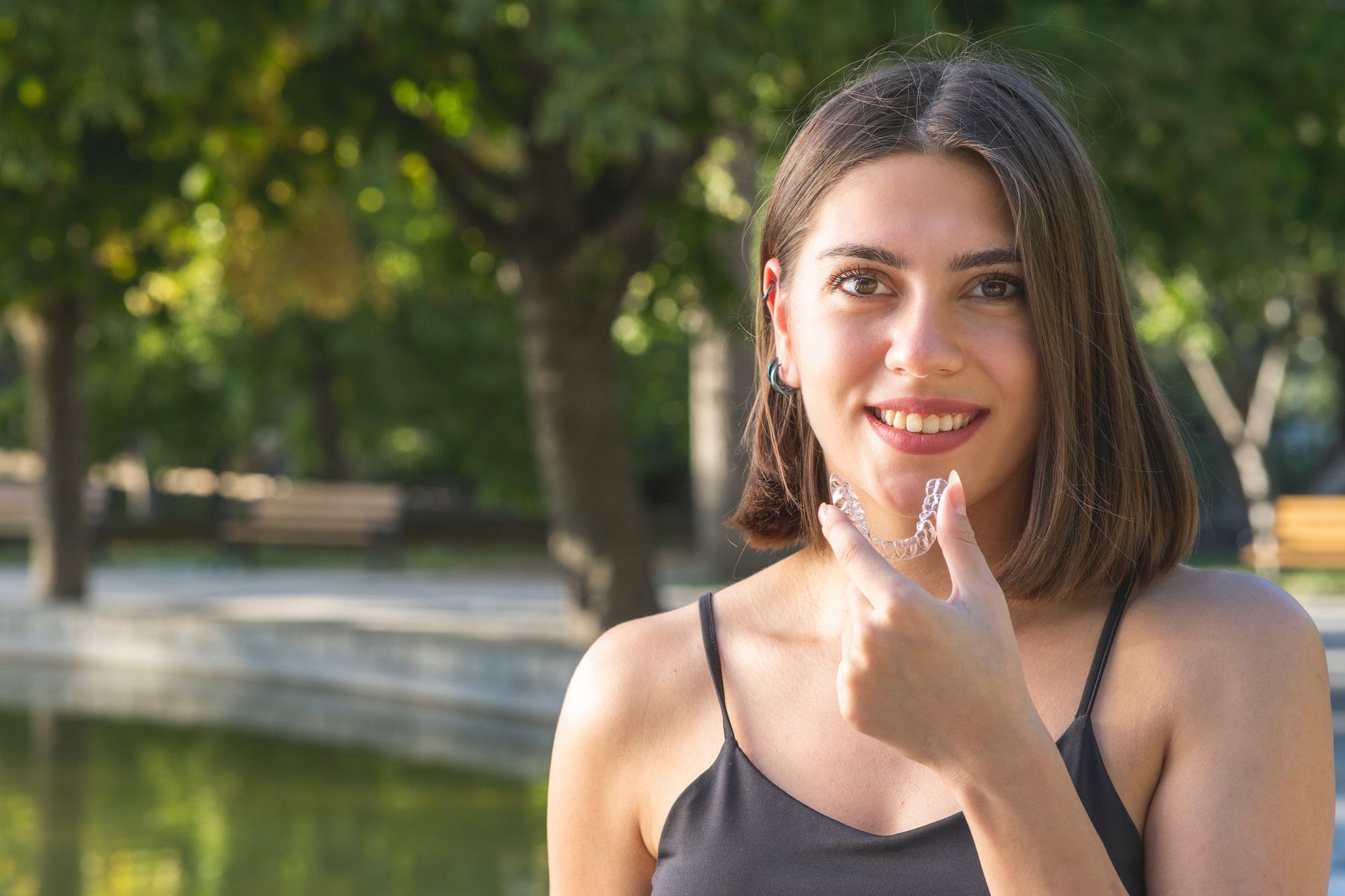 Smiling woman in a black tank top standing outdoors in a park, holding an clear aligner.