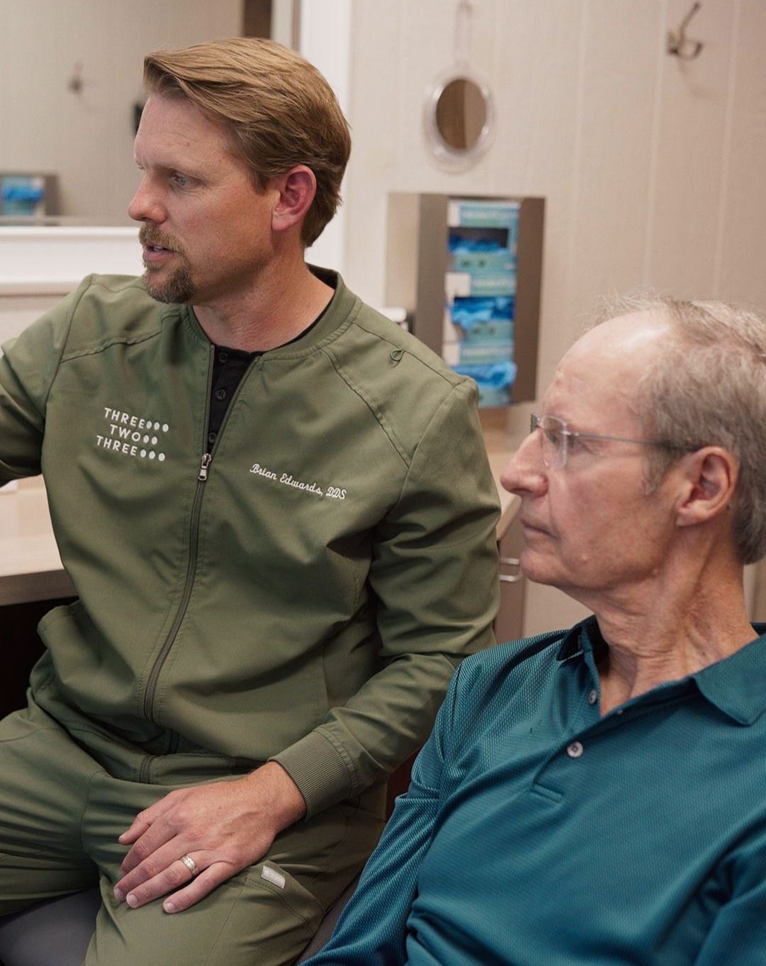 Two men seated in a room, one in a green jacket and one in a teal shirt, listening attentively.