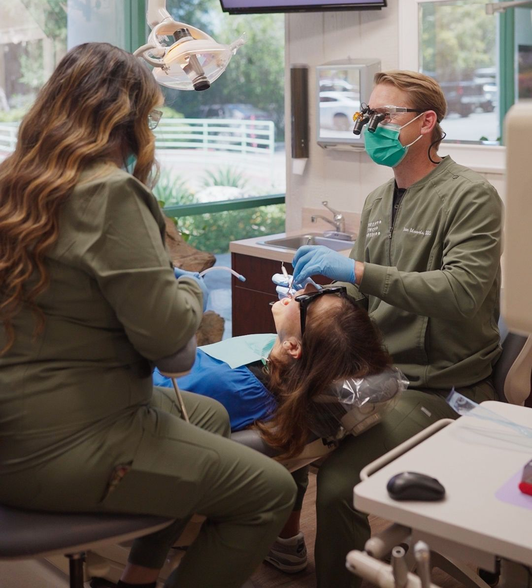 Dentist treating a patient in a dental chair, with a masked assistant nearby in a bright clinic.