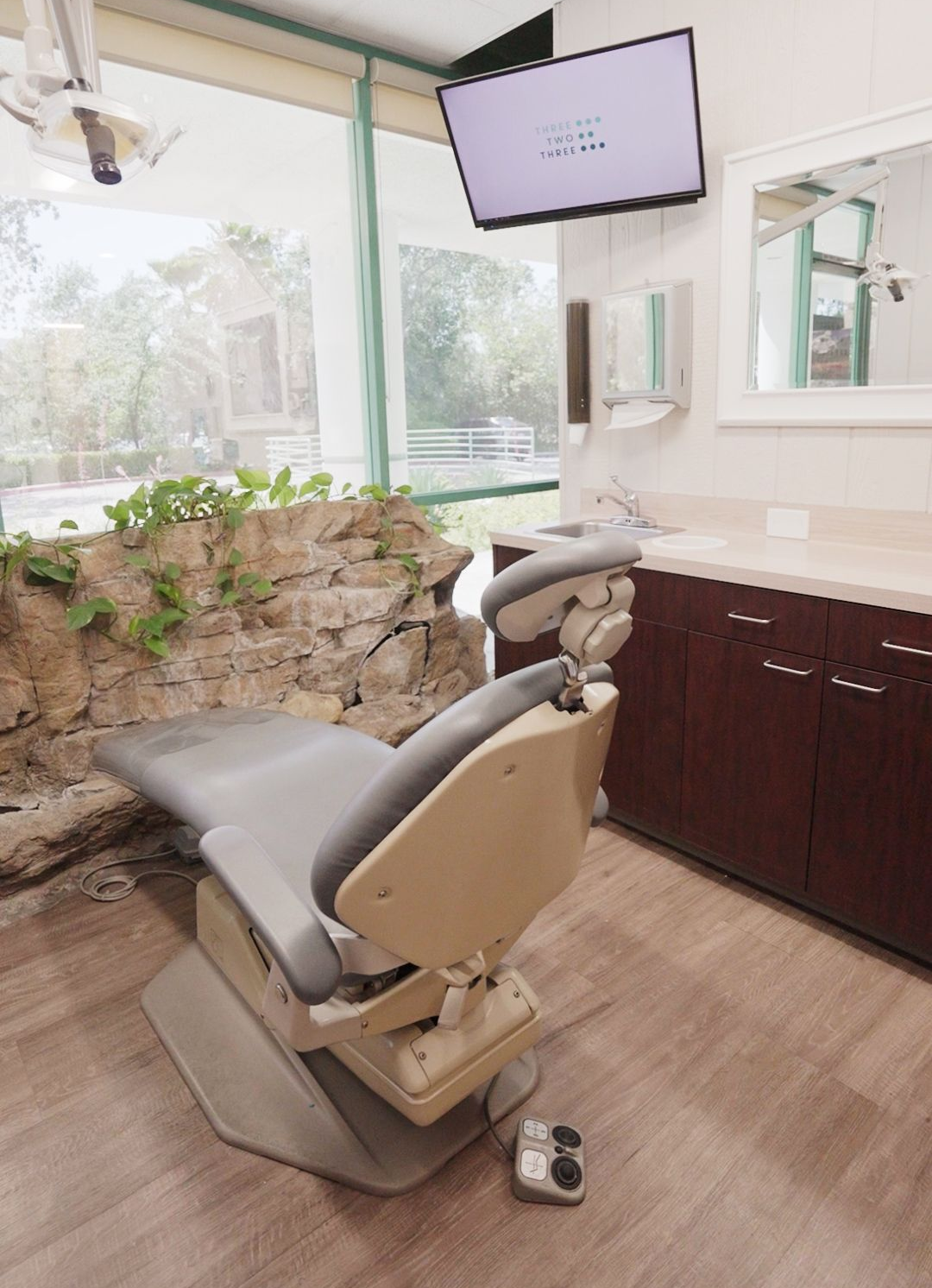 Dental exam chair in a bright clinic, with monitor, sink, and large windows.
