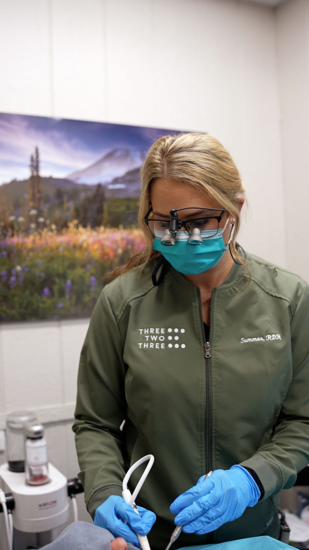 Dental professional in green scrubs, mask, and gloves preparing tools in a clinic room.