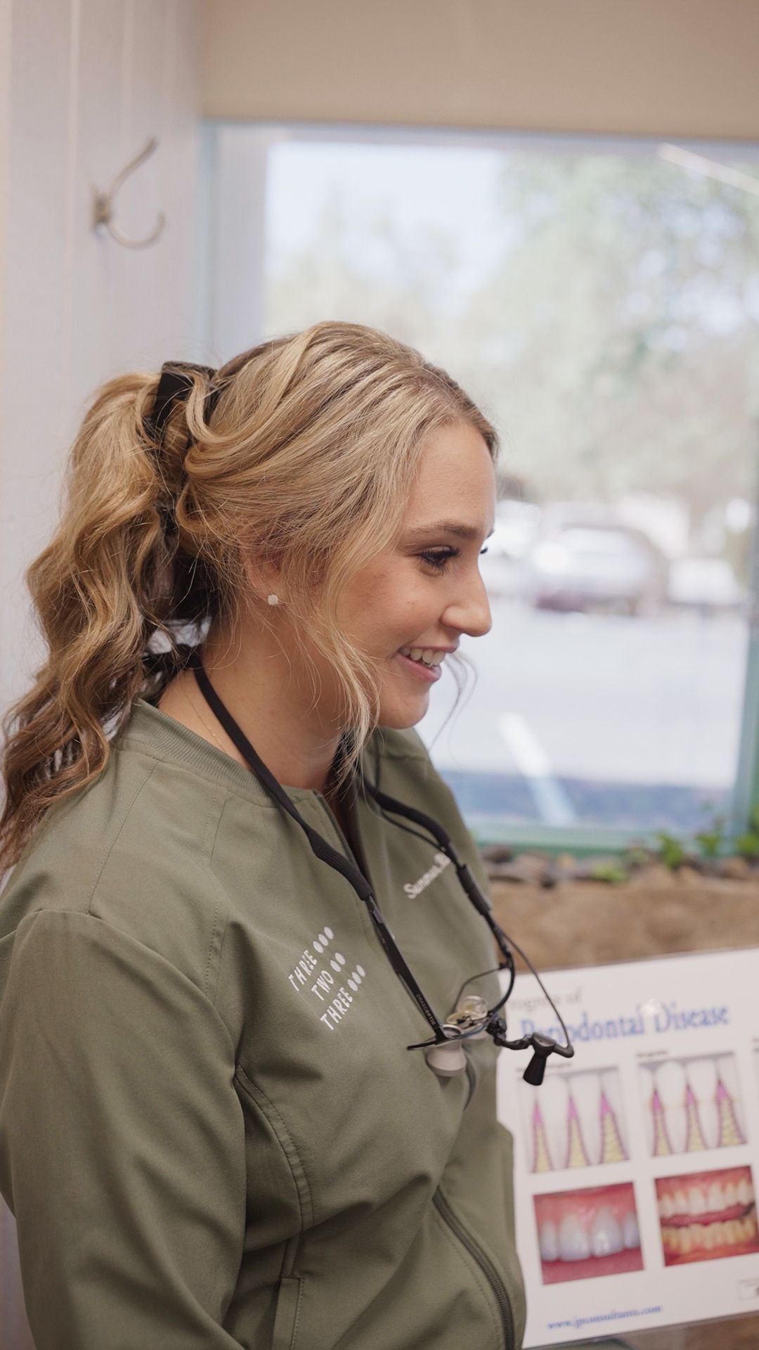 Woman in olive-green scrubs smiling in a clinic, wearing a black lanyard and facing a wall chart.