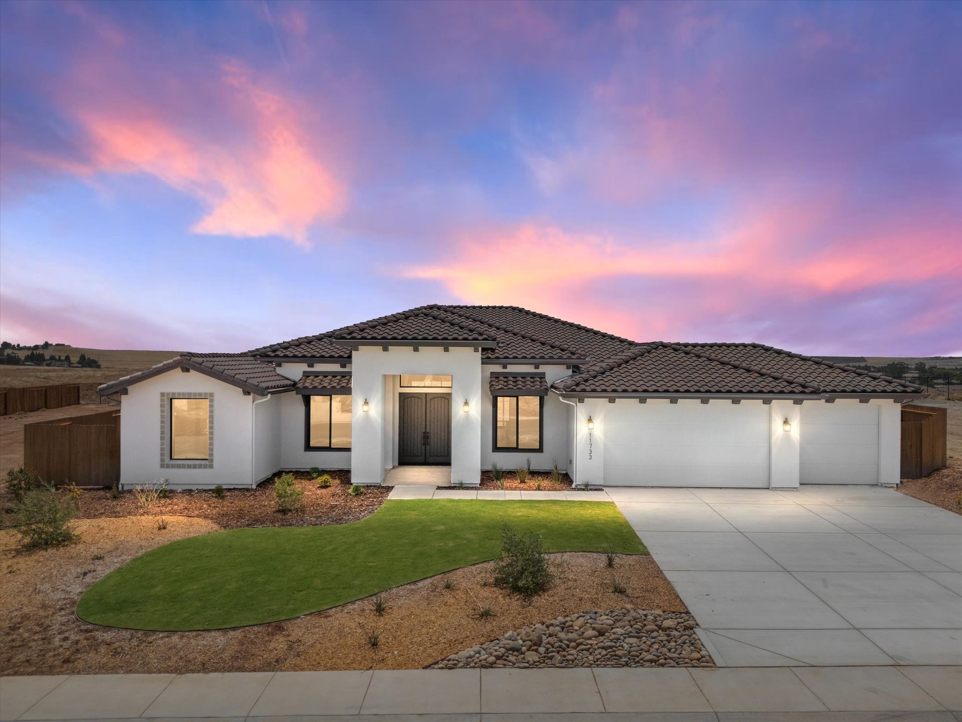 Modern white house with a tile roof, driveway, and landscaping under a colorful sunset.
