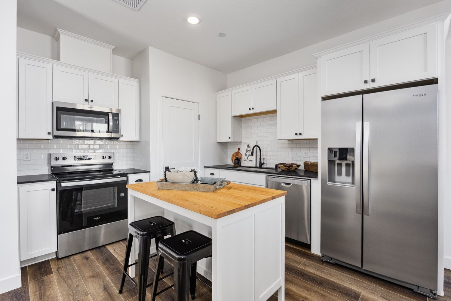 Bright white kitchen with stainless steel appliances, wooden island, and two black stools.