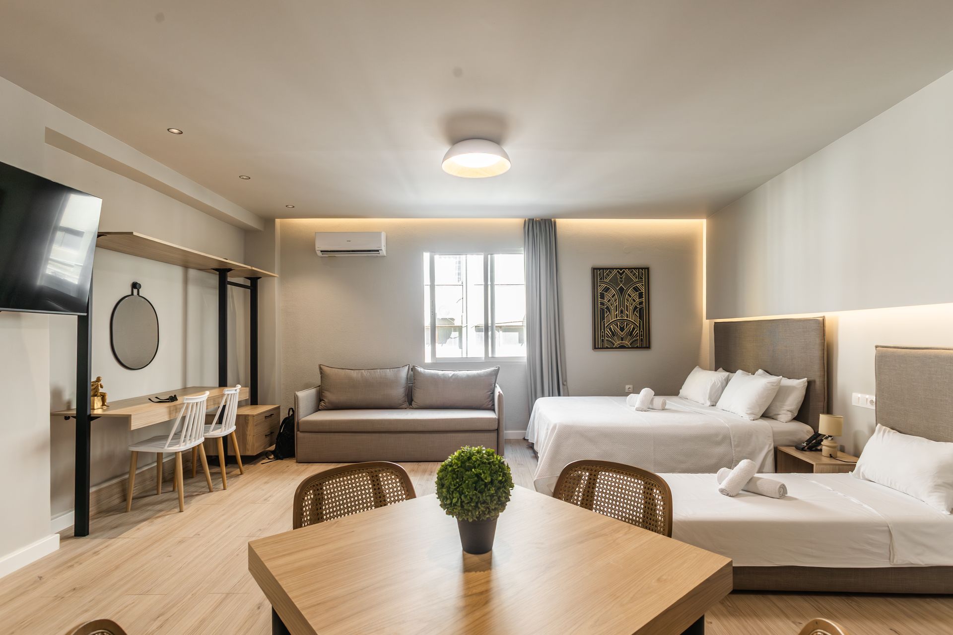 Spacious hotel room with three beds, a sofa, and a table. Bright, neutral colors.