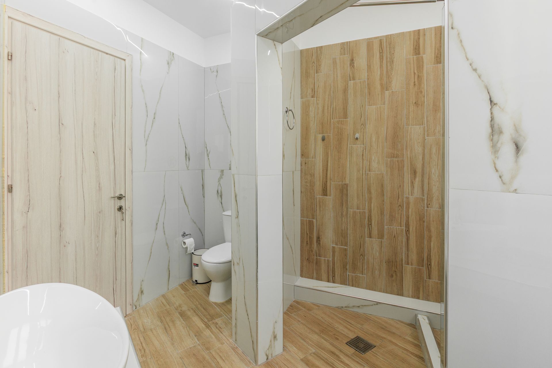 Bathroom with white marble walls, wooden shower wall, toilet, and a closed wooden door.
