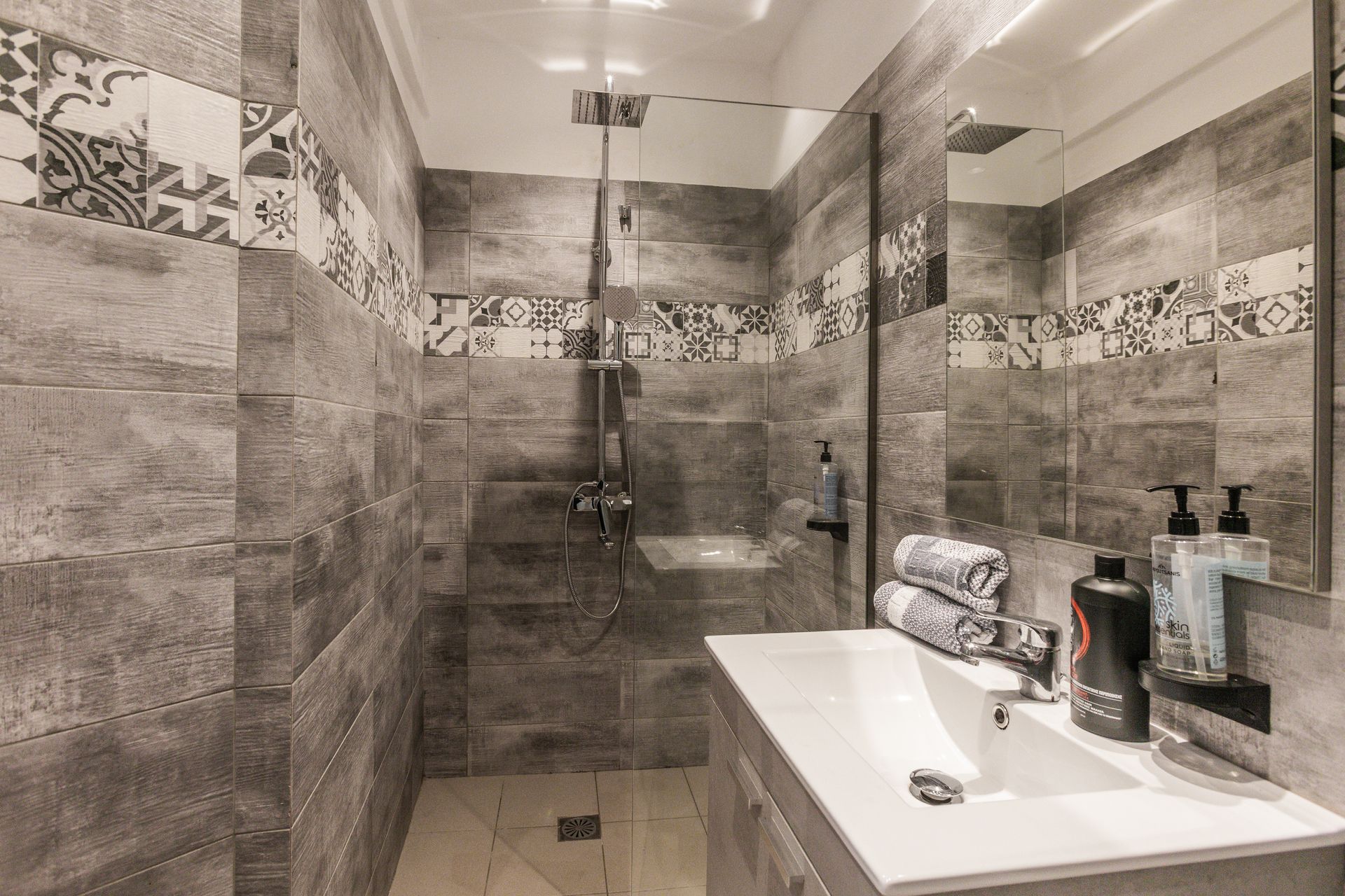 Gray-tiled bathroom with shower, sink, and mirror. Decorative tiles border the walls. Two folded towels and toiletries are on the counter.