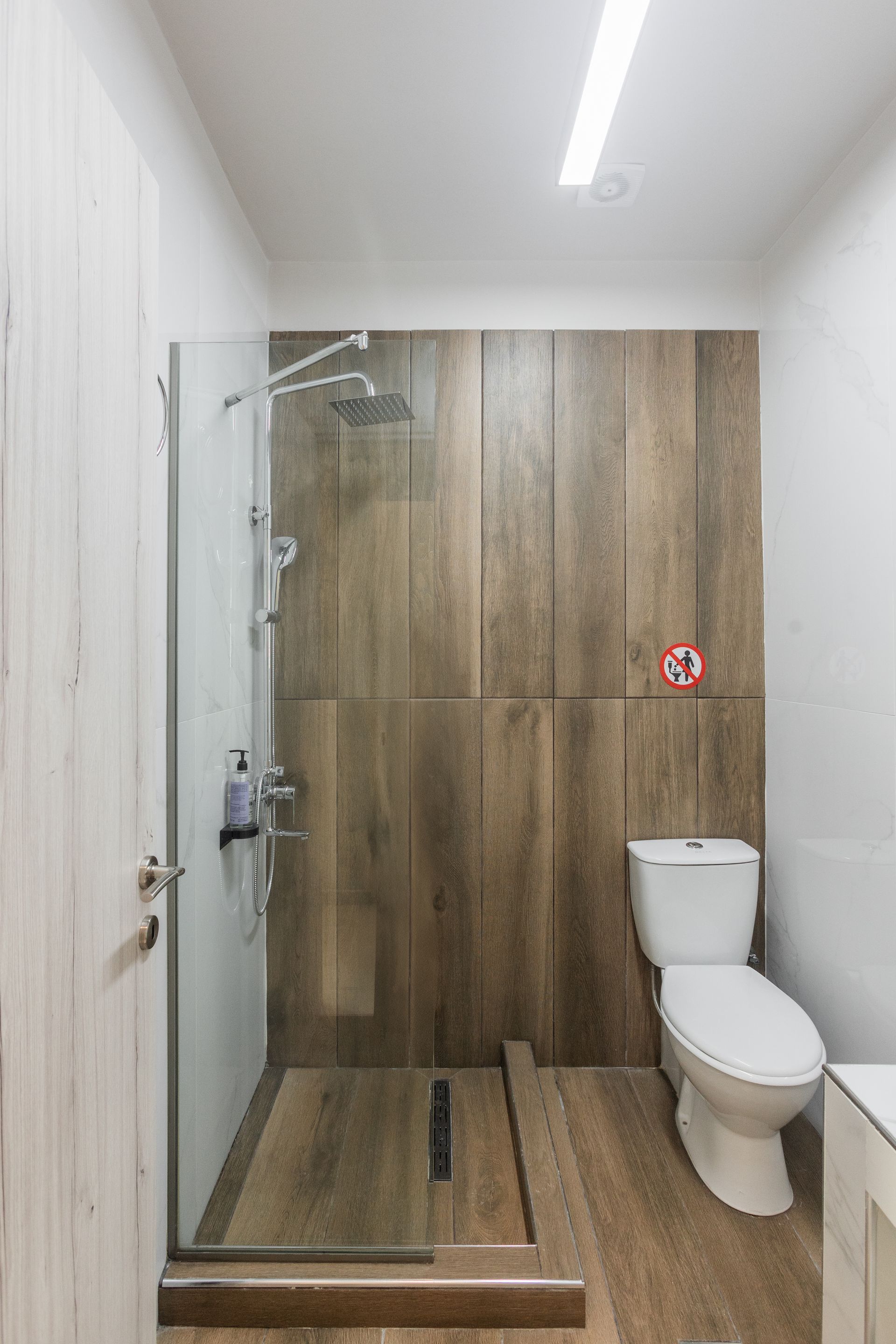 Bathroom with a wood-paneled shower, glass door, white toilet, and wooden floor.