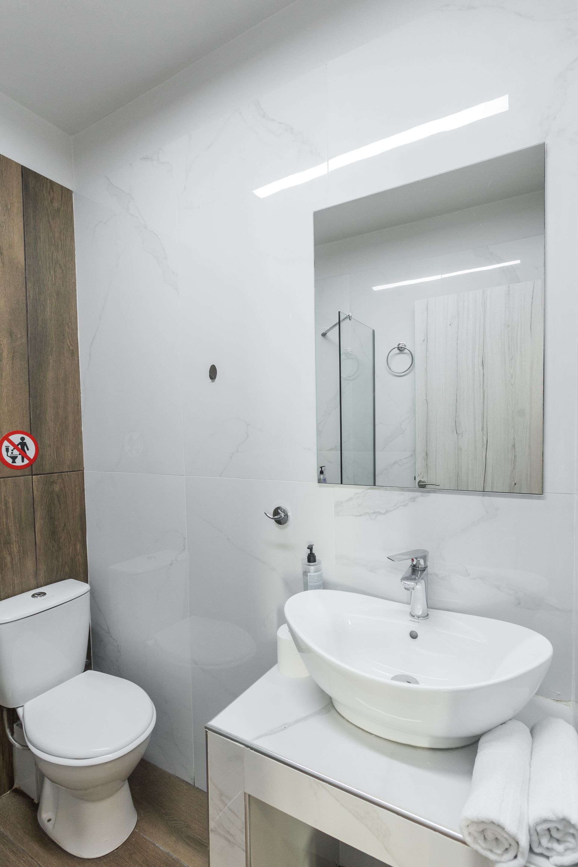 White bathroom with toilet, sink, mirror, and shower. Wooden accent wall.