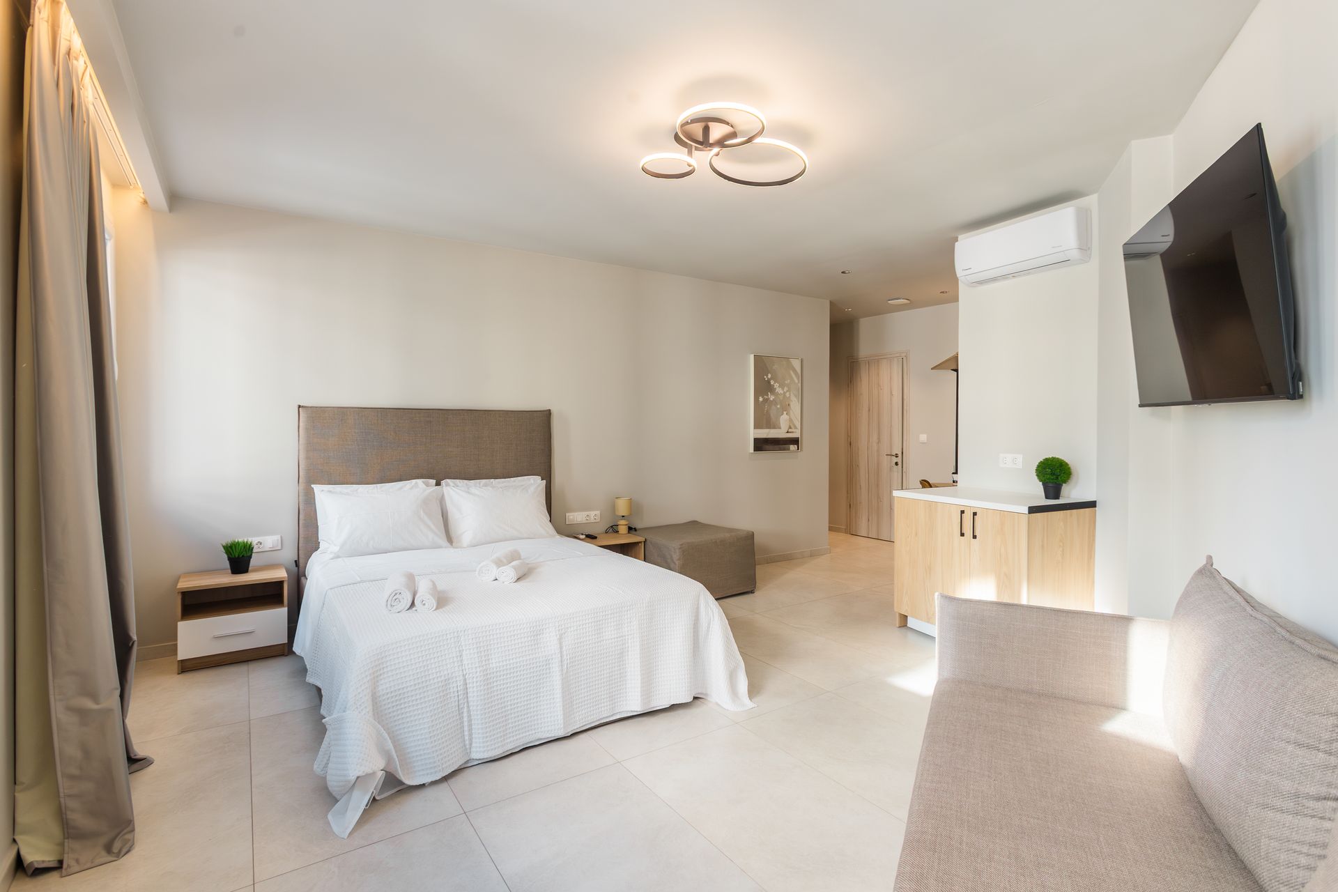 Bright hotel room with bed, sofa, cabinet, TV, and decorative plants.