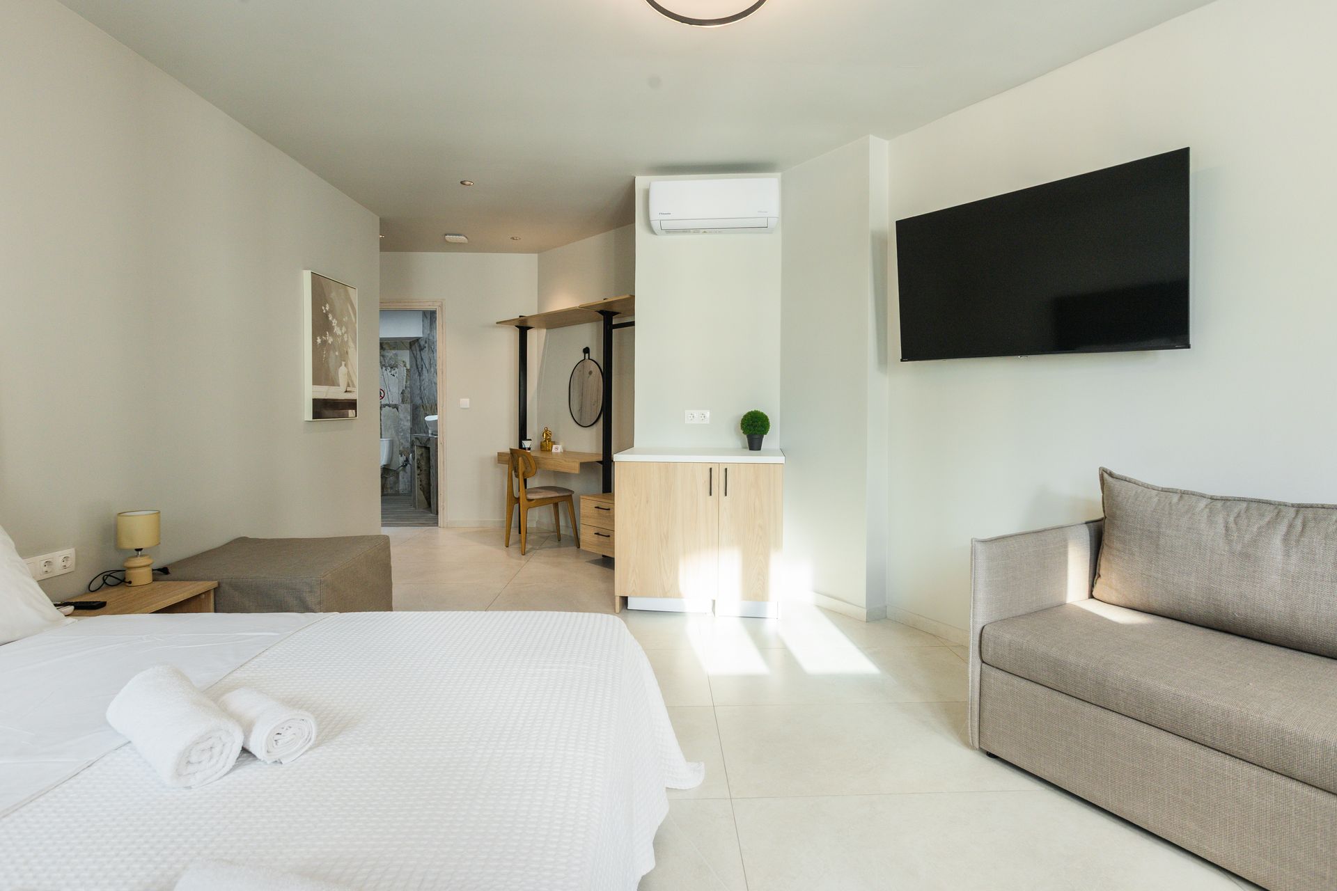 Bright modern studio apartment with bed, sofa, TV, small kitchenette, and bathroom visible in the background.