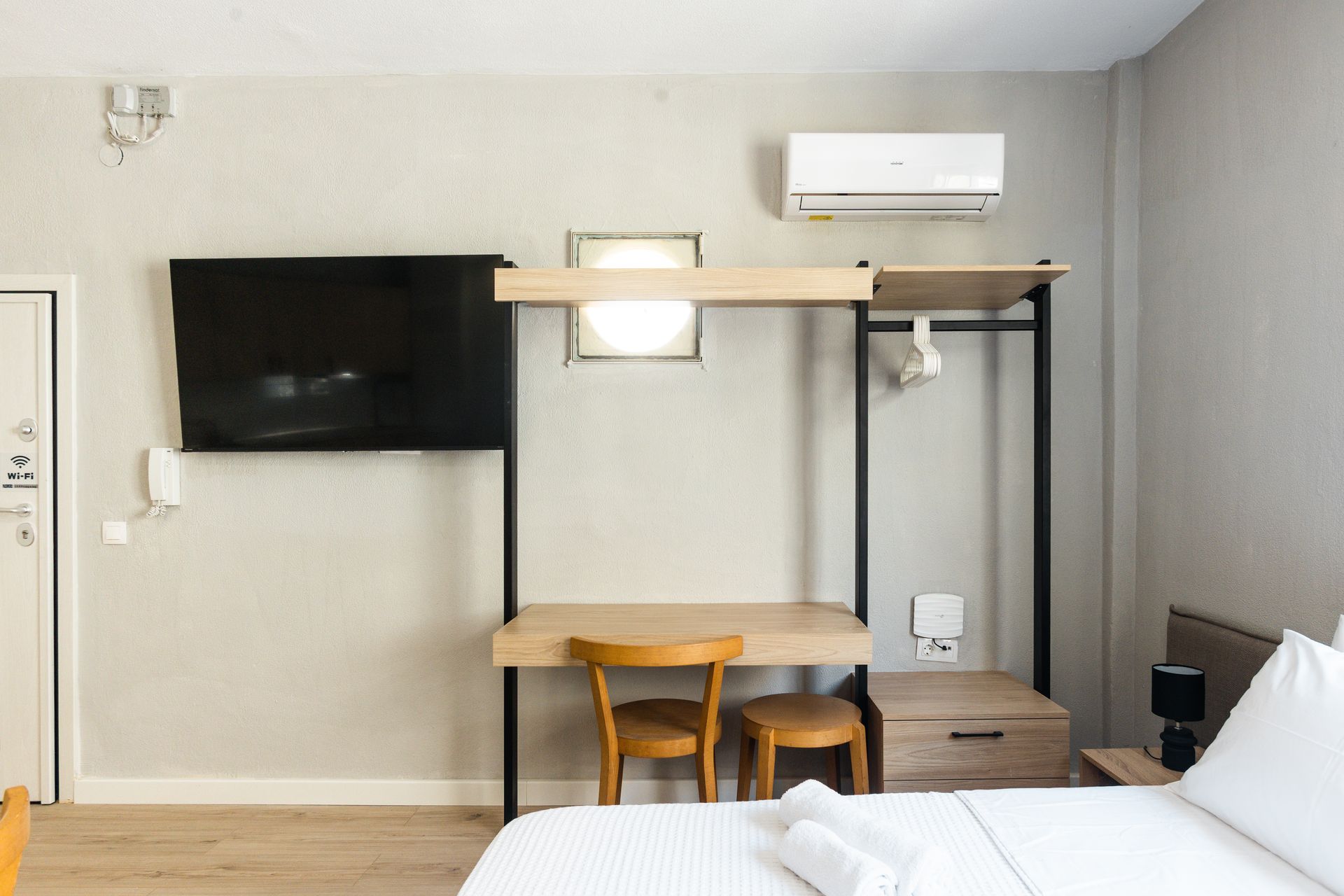 Bedroom with desk, TV, air conditioner, and bed. Light wood furniture against a light gray wall.
