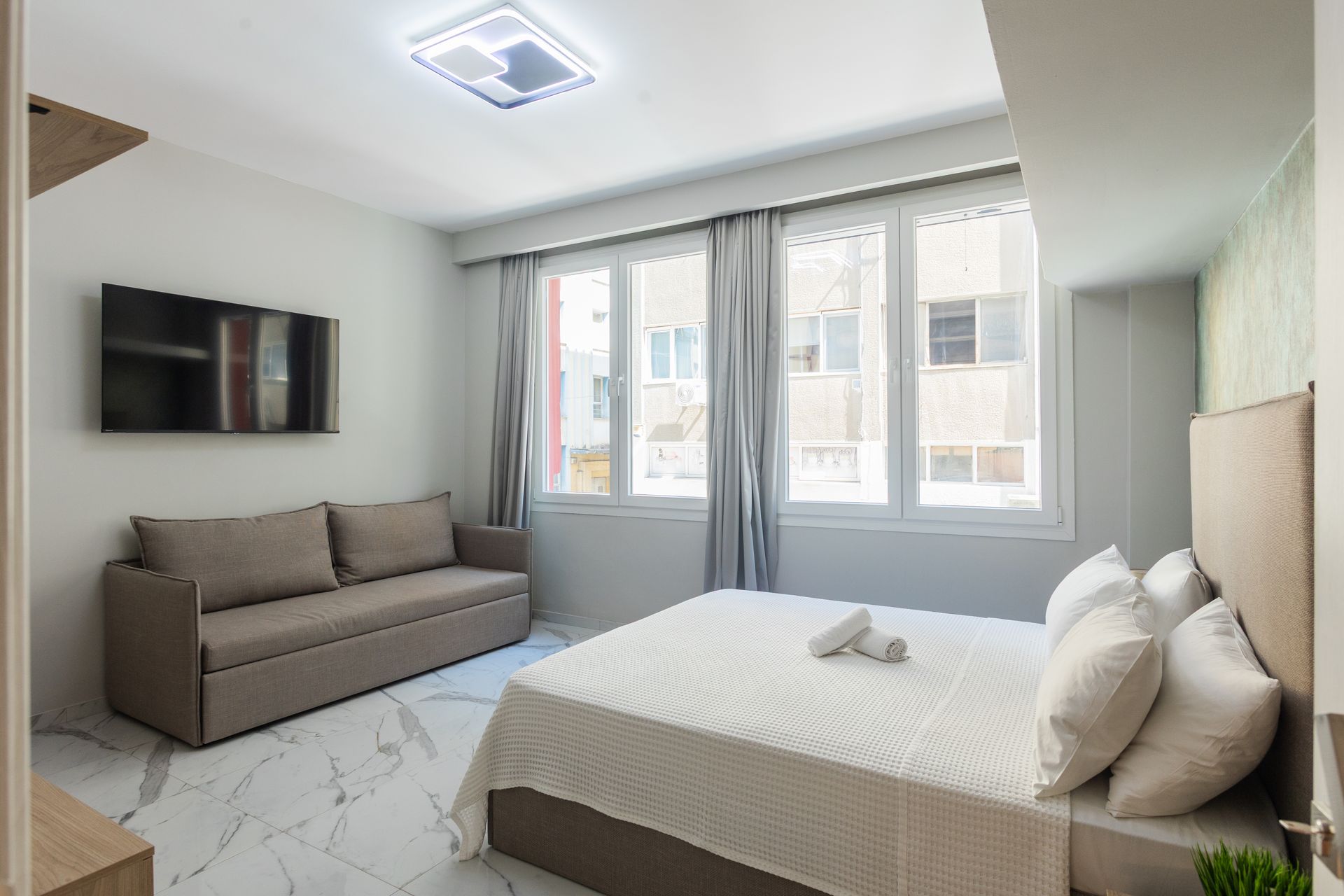 Bedroom with bed, sofa, large windows, TV on wall; white and gray tones.
