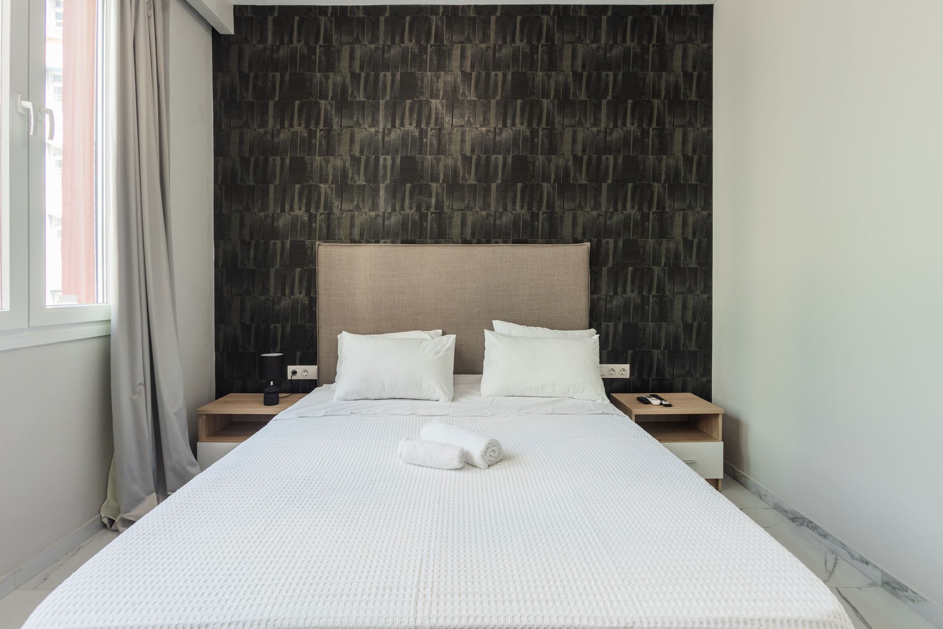Bedroom with double bed, bedside tables, patterned wallpaper, and window with curtains.