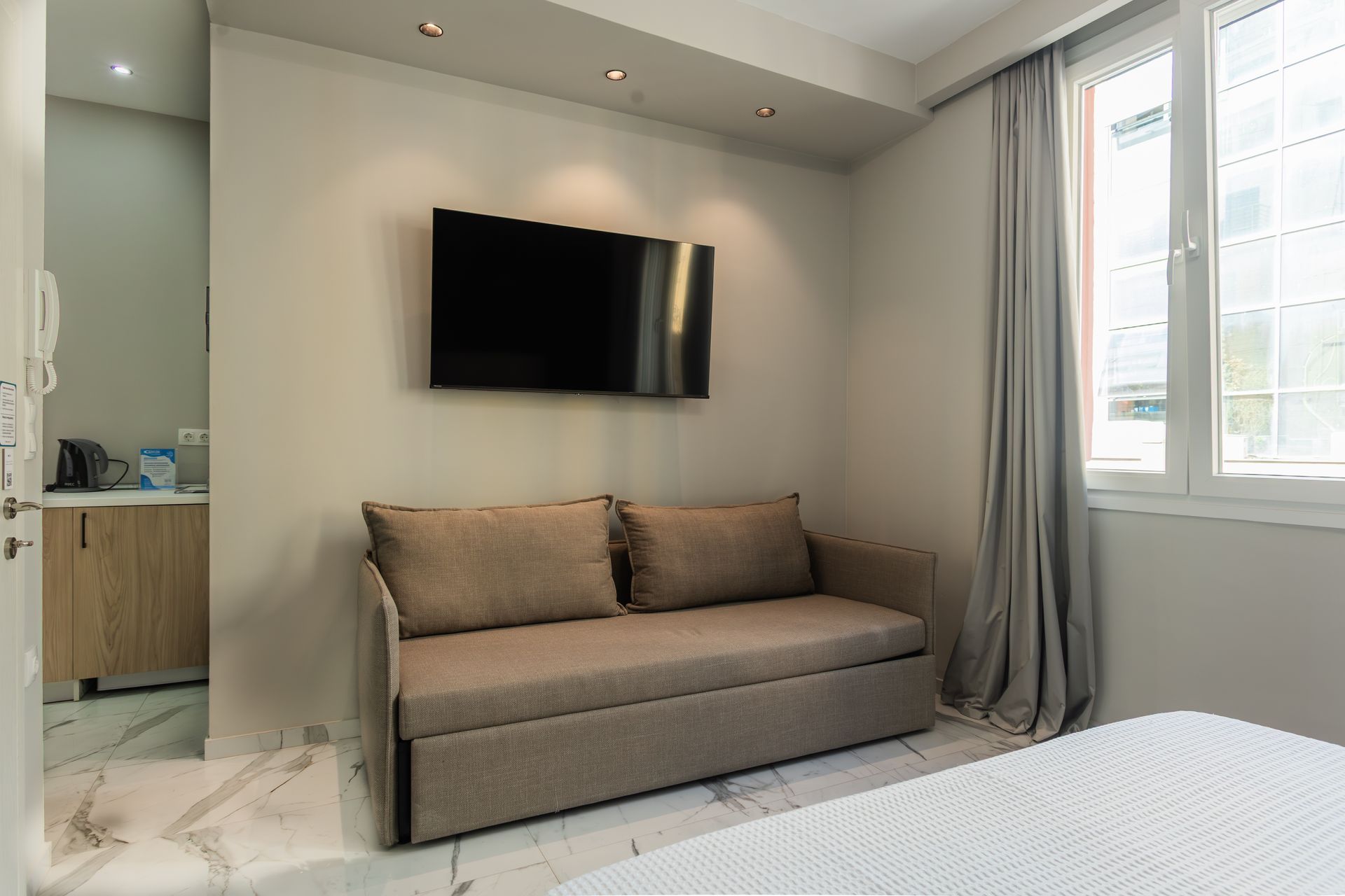 Small room with sofa, TV, and window. Light walls, gray curtains, and marble floors.