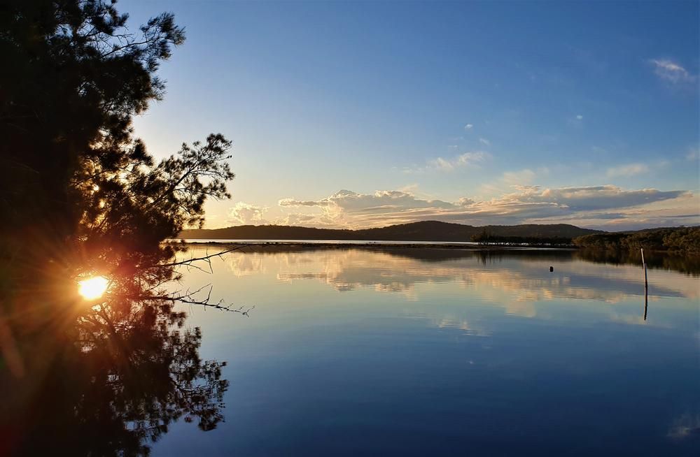 The Sun is Setting Over a Lake With Trees in the Foreground — Roofing Contractor in Laurieton, NSW