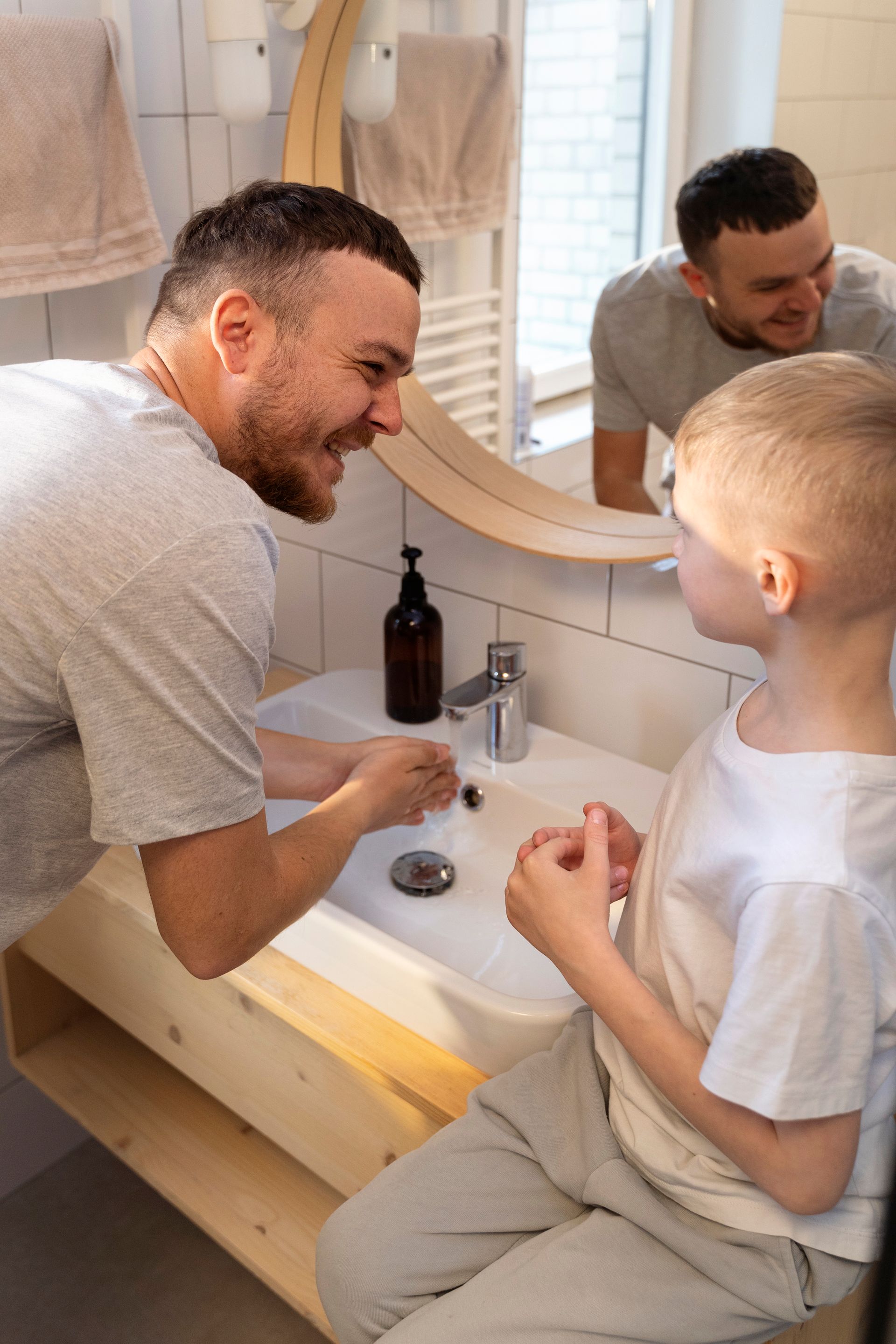 Man brushing a boy’s teeth at a bathroom sink, both smiling and facing the mirror.